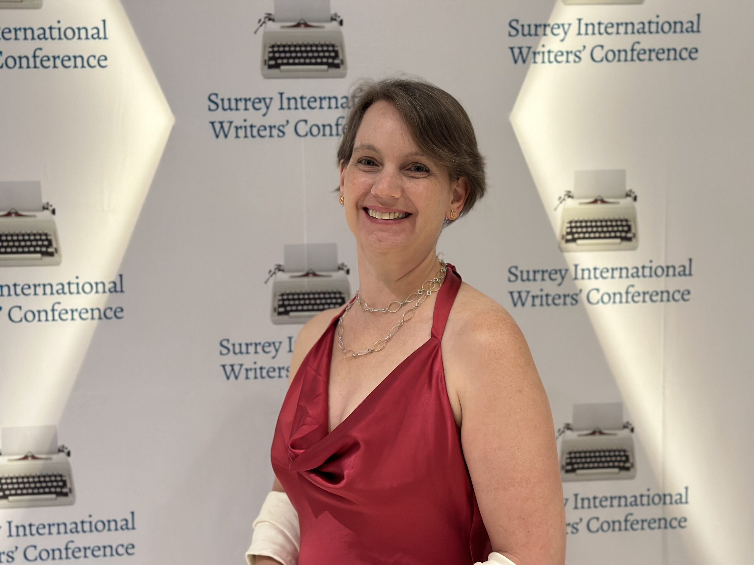 White person with short-ish brown hair in a red dress against a background with typwriters on it and Surrey International Writers Conference